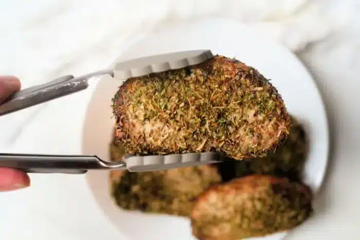 A pair of tongs holds a herb-crusted pork steak over a white plate with more air fryer pork steaks in the background. The coating appears to consist of various green herbs.