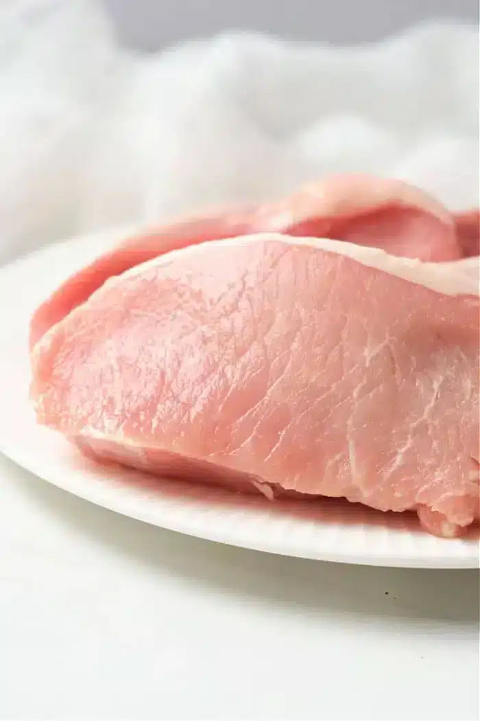 Close-up of raw, boneless pork chops prepared for an air fryer on a white plate. The fresh, pink meat boasts visible marbling against a softly blurred, white background.