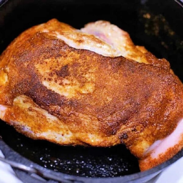 A piece of perfectly seasoned pork roast is cooking in a black cast iron skillet. The meat boasts a golden-brown crust, indicating it's being seared or roasted to perfection. While it may remind you of a good dutch oven dish, this culinary masterpiece rests confidently on the stove's cooking surface.