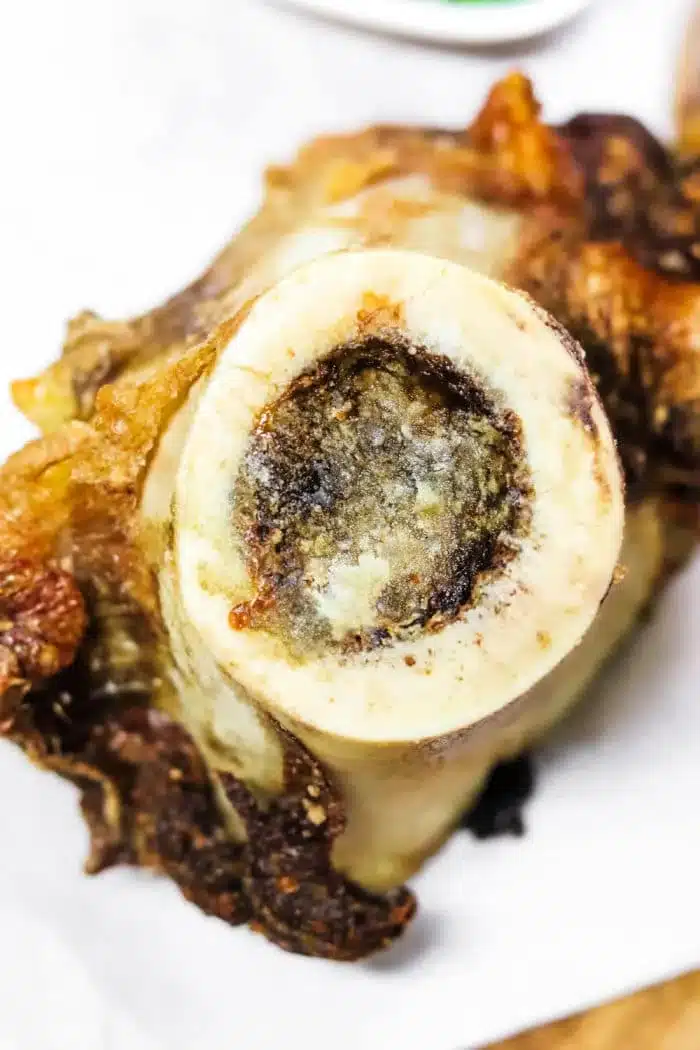 Close-up of a roasted bone with exposed bone marrow at the center, air fryer crisping its outer edges to perfection. The background is blurred, set against a white surface beneath the bone.