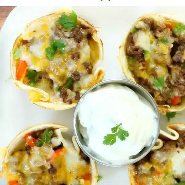 Four steak fajita cups overflowing with ground meat, melted cheese, and vegetables are artfully arranged on a plate. In the center, a small dish of sour cream garnished with a cilantro leaf waits to be savored. Above reads: Fajita Cups, the best little appetizer ever!