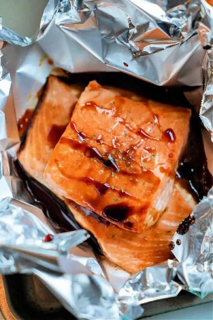 Cooked oven-baked salmon fillets glazed with a rich teriyaki sauce, garnished with herbs, are placed on a sheet of aluminum foil. The foil surrounds the fish, creating a partially open cooking pouch.