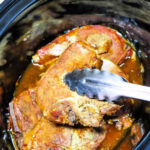 Close-up of Slow Cooker Ranch Pork Chops in a cooker, being lifted with metal tongs.