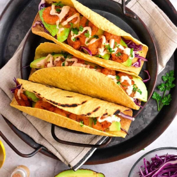 Three tacos with chicken, avocado, cabbage, and sauce sit invitingly on a plate. Beside them is a fresh avocado half and vibrant purple cabbage. These delicious tacos feature perfectly crispy taco shells made in an air fryer for the ultimate crunch experience.