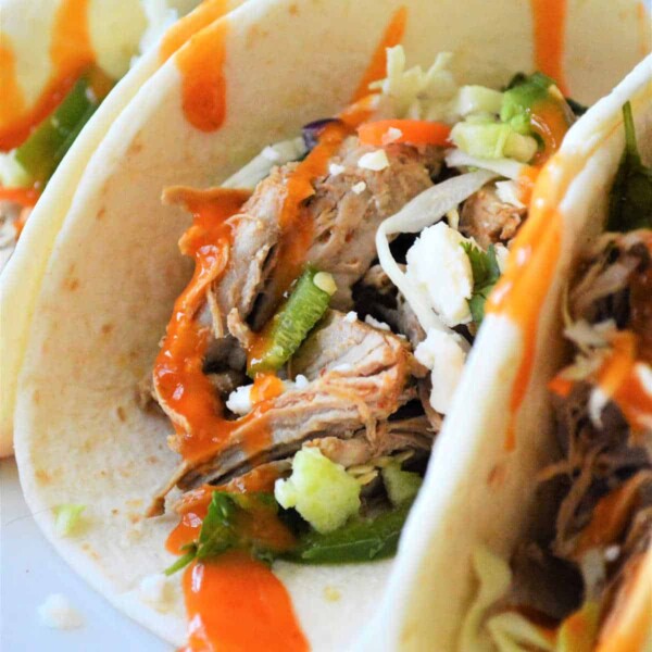 Close-up of instant pot pork tacos brimming with shredded meat, chopped vegetables, cheese, and a drizzle of hot sauce.