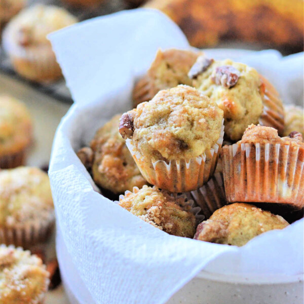 A bowl of mini banana nut muffins lined with a white napkin, with more delicious mini banana muffins and ripe bananas in the background.