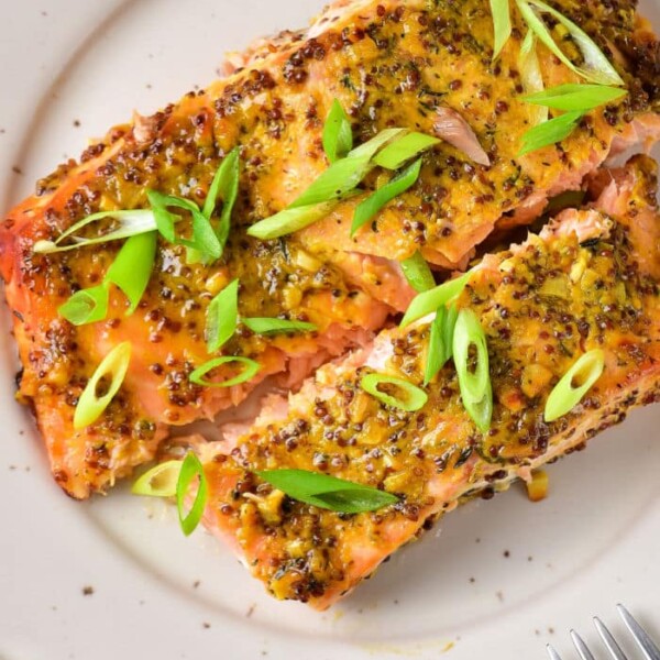 Plate with two slices of salmon topped with a rich mustard sauce and garnished with sliced green onions. A fork is on the side.