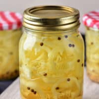 A jar of pickled banana peppers takes center stage, showcasing a metal lid and visible peppercorns. In the blurred background, two more jars with checkered lids hint at an assortment of pickled fruits and vegetables, all elegantly displayed on a wooden surface.