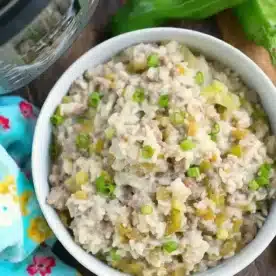 A bowl of creamy instant pot sausage rice casserole garnished with chopped green onions sits on a wooden surface next to whole green peppers and a colorful floral kitchen towel. An Instant Pot is partially visible in the corner, hinting at the delicious simplicity of this comforting dish.