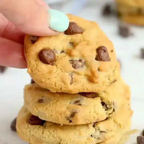 A hand holding a freshly baked chocolate chip cookie above a stack of three similar air fryer cookies. Chocolate chips are scattered on a white surface around the cookies, enhancing their delicious appeal.