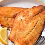Golden-brown grilled fish fillets, cooked to perfection using air fryer techniques, rest on a plate with lemon wedges and a fork for the perfect healthy recipe.