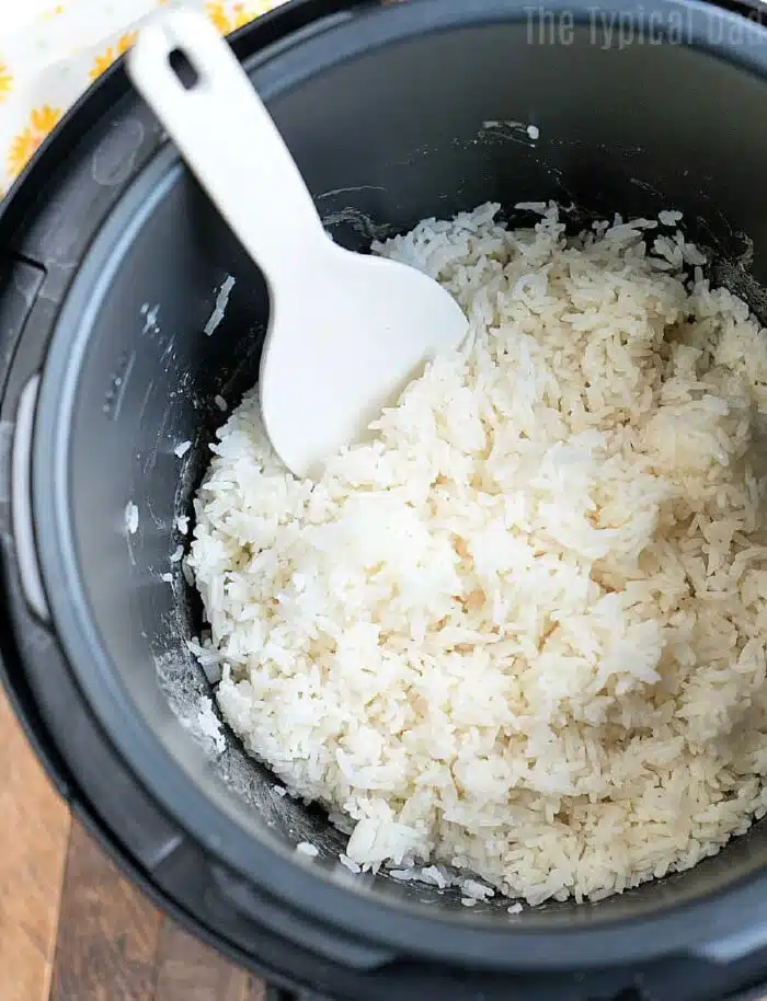 A Crockpot Express is filled with perfectly cooked white rice, and a white plastic rice paddle rests atop the fluffy grains. The cooker sits on a warm wooden surface, ready to serve.