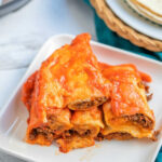 A plate of cheesy beef enchiladas, made perfectly in an instant pot, is artfully stacked on a white square plate with a hint of tortillas in the background.