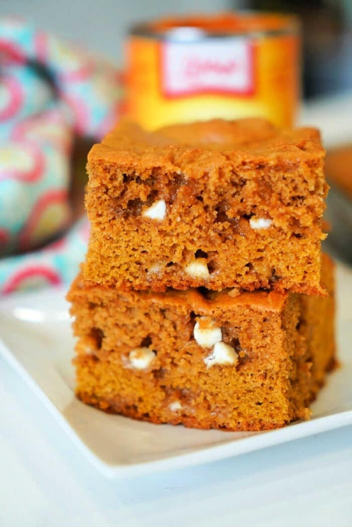 Two delicious pumpkin blondies with white chocolate chips are stacked invitingly on a pristine white plate, set against a vibrant, colorful cloth in the background.