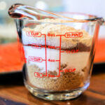 A clear measuring cup filled with salt and brown sugar sits on a wooden surface, resembling the perfect blend for a salmon dry rub.