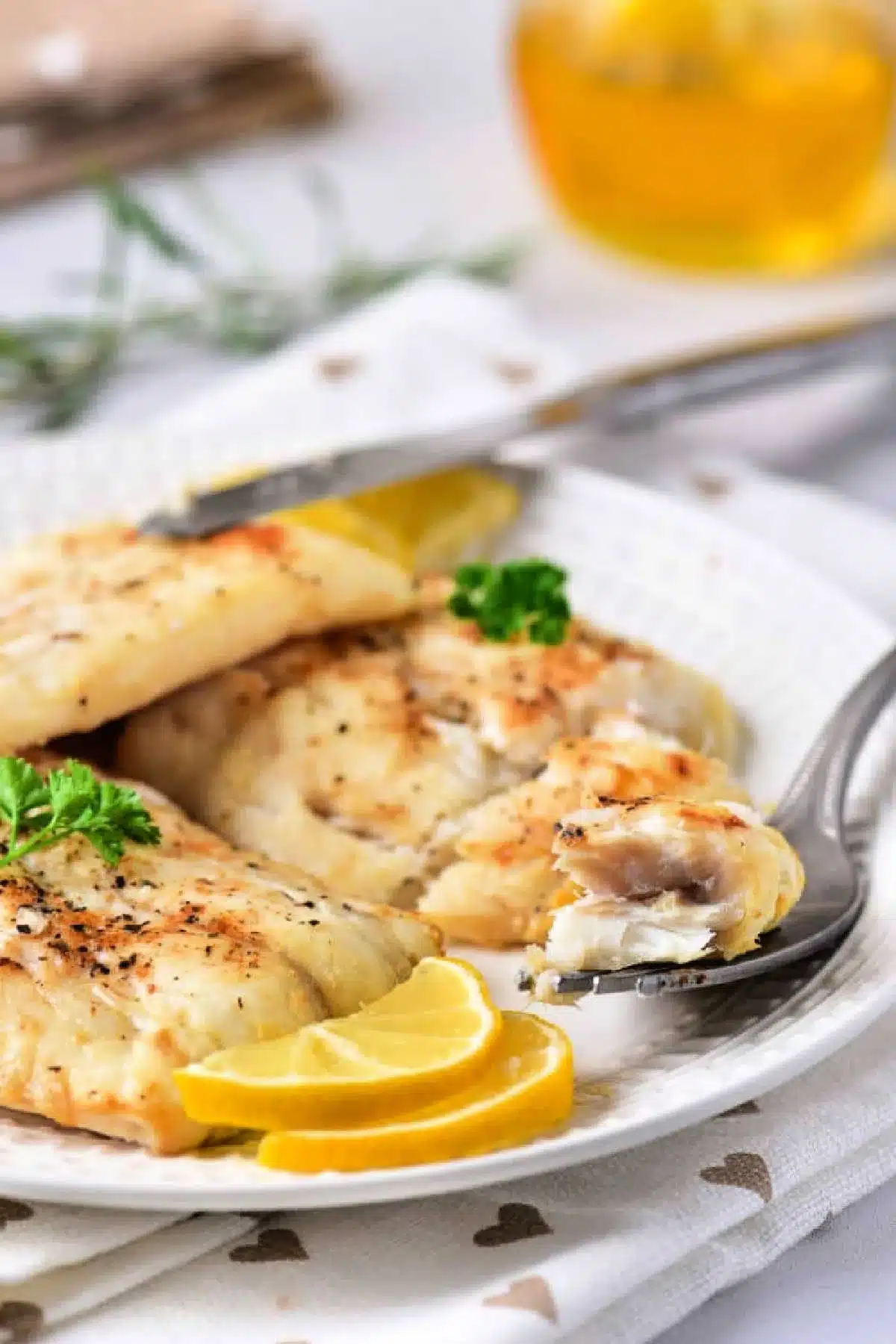 A white plate features air-fried fish fillets garnished with lemon slices and a sprig of parsley. A fork holds a piece of the flaky delight, while a blurred glass with a yellow drink sits in the background. The tablecloth is white with heart patterns, emphasizing this delicious twist on frozen cod.