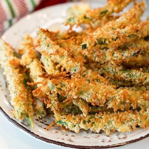 A plate of golden-brown, crispy air fryer green bean fries. They are sprinkled with crumbs and arranged in a heap on a rustic white plate, creating an appetizing display.