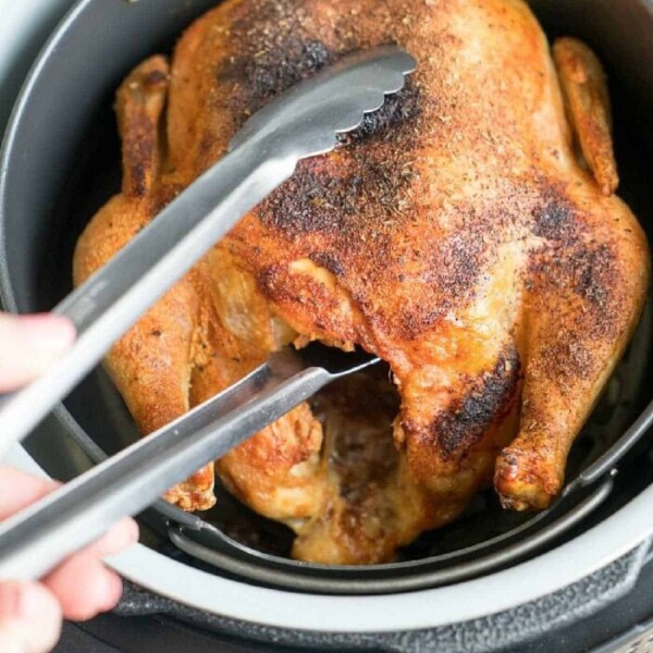 A delicious air fryer whole chicken is inside an air fryer. A pair of metal tongs is shown grabbing the chicken. The bird boasts a browned, crispy skin with visible seasoning, making it irresistibly appealing and packed with flavor.