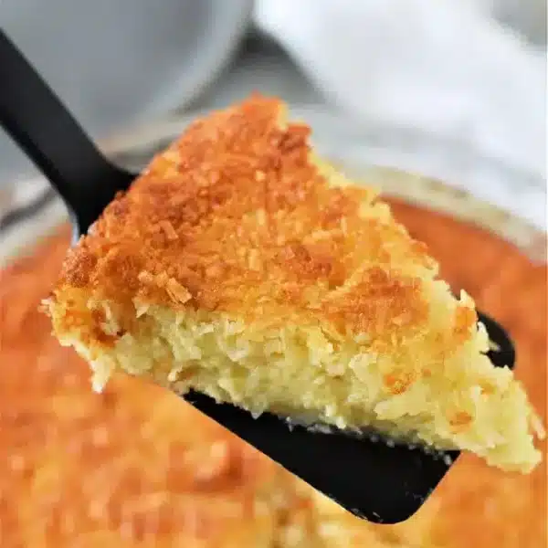 A slice of golden-brown coconut pie, akin to a Bisquick Impossible Pie recipe, is being lifted with a black spatula from the round dish. The pie boasts a crispy top and moist interior, while a white cloth peeks in the background.