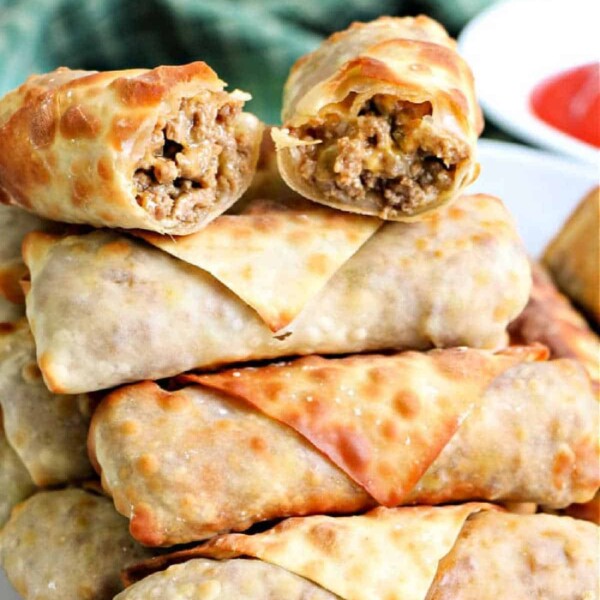 A plate stacked with several golden-brown ground beef egg rolls. The top egg roll is halved, revealing a delicious filling of savory meat and vegetables. A green cloth is partially visible in the background.