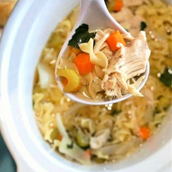 A spoonful of chicken soup in the crockpot is held above a white bowl, revealing ingredients like noodles, carrots, chicken, and greens.