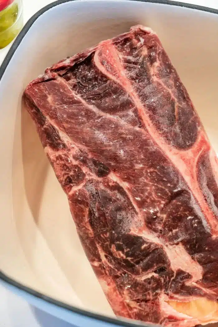 A raw beef chuck roast in a white oblong dish, ready to become tender shredded beef from frozen. The meat has visible marbling and fat along the edges.