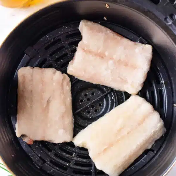 Three frozen cod fillets are placed in an air fryer basket. Nearby, there is a small bowl of spices, a glass bottle of oil, rosemary sprigs, and lemon slices on a light-colored surface.