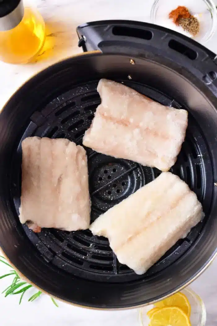 Three frozen cod fillets are placed in an air fryer basket. Nearby, there is a small bowl of spices, a glass bottle of oil, rosemary sprigs, and lemon slices on a light-colored surface.