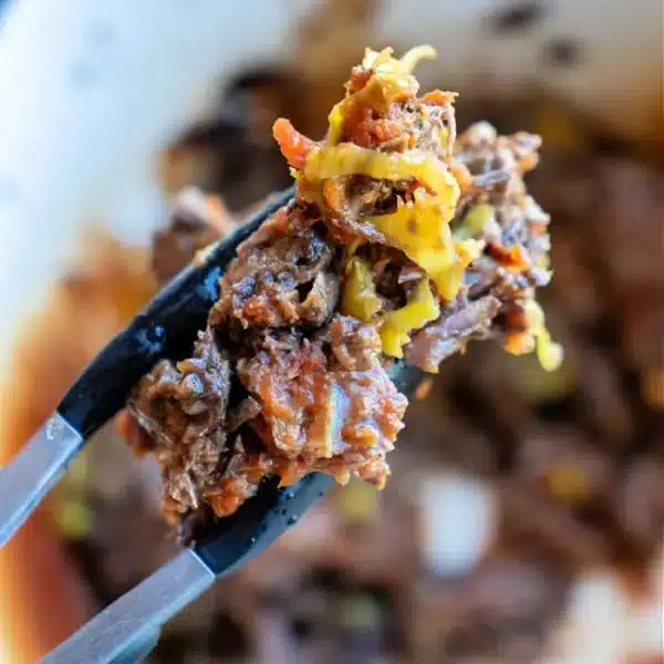 Close-up of a fork holding a piece of cooked shredded beef from frozen, mixed with melted cheese and diced vegetables, with more of the savory dish blurred in the background.