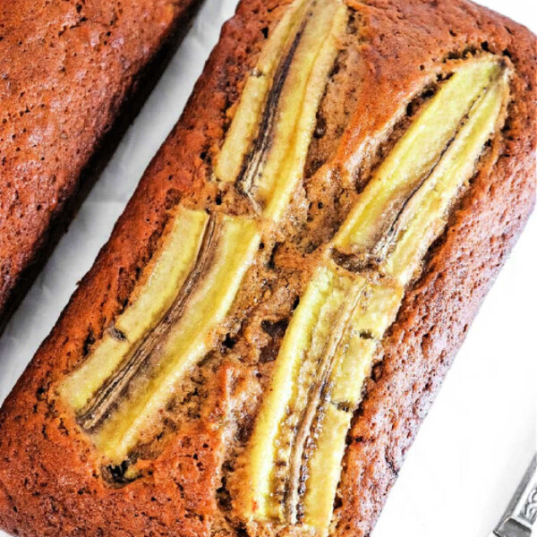 A loaf of ginger banana bread with sliced bananas on top rests elegantly on a pristine white surface.