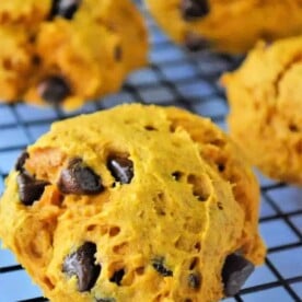 Close-up of pumpkin cookies with cake mix and chocolate chips on a cooling rack. The vibrant orange cookies speckled with rich chocolate are set against a blurred background, showcasing even more delicious treats.