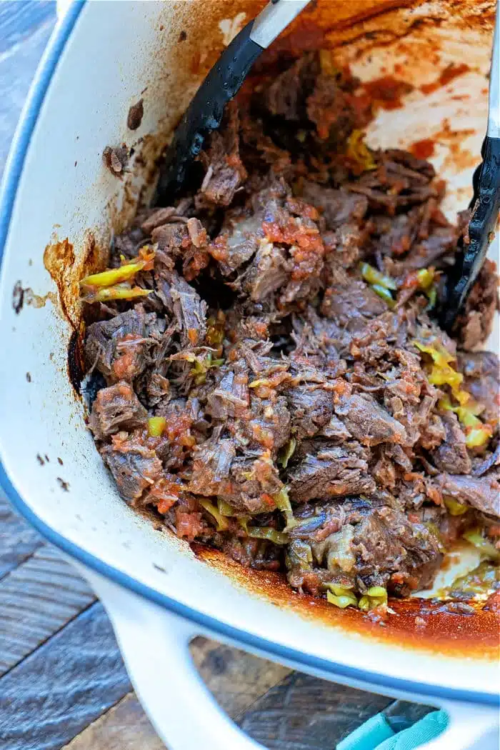 A white pot filled with shredded beef, made from frozen, includes visible chunks of vegetables like tomatoes and green peppers. Black tongs rest inside the pot, and sauce residue clings to the sides. The pot is set upon a wooden surface.