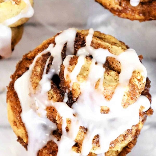 A close-up of a cinnamon roll muffin with cream cheese icing drizzled on top. The muffin has visible swirls and rests on a light, marble-colored surface. Several wedges of additional muffins are partially visible around the main roll.