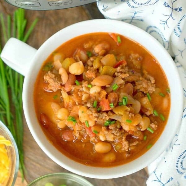 A bowl of pressure cooker chili with dry beans, meat, and vegetables, garnished with chives, sits enticingly next to fresh onions and cheese.