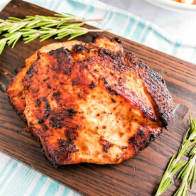 Grilled chicken breast with a crispy exterior rests on a wooden board, adorned with sprigs of rosemary. In the background, an air fryer turkey breast and a bowl of roasted potatoes sit charmingly on a striped cloth.