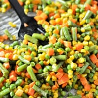 A close-up of roasting mixed vegetables, including green beans, corn, peas, and carrots on a baking sheet, with a black spoon resting on the side.