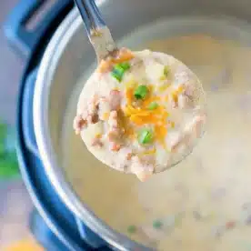 A ladle filled with creamy, instant pot cheeseburger soup brimming with chopped green onions, shredded cheese, and small meat chunks hovers above an Instant Pot filled with the same delicious soup. The edge of a cutting board showcasing extra toppings peeks in the background.