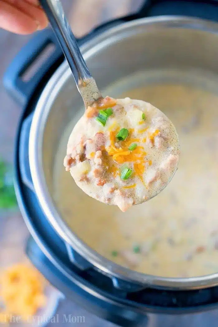 A ladle filled with creamy, instant pot cheeseburger soup brimming with chopped green onions, shredded cheese, and small meat chunks hovers above an Instant Pot filled with the same delicious soup. The edge of a cutting board showcasing extra toppings peeks in the background.