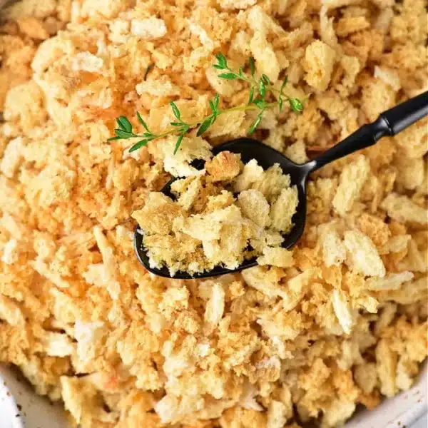 A bowl filled with golden Italian panko bread crumbs, with a black spoon resting on top. A small sprig of green herbs is placed on the spoon, providing a hint of color contrast.