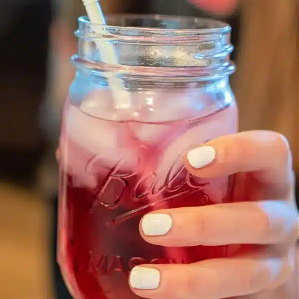 A hand with white-painted nails holds a mason jar filled with a pink iced Lotus drink and a white straw. The jar is half-full, radiating refreshing vibes against the softly blurred background.