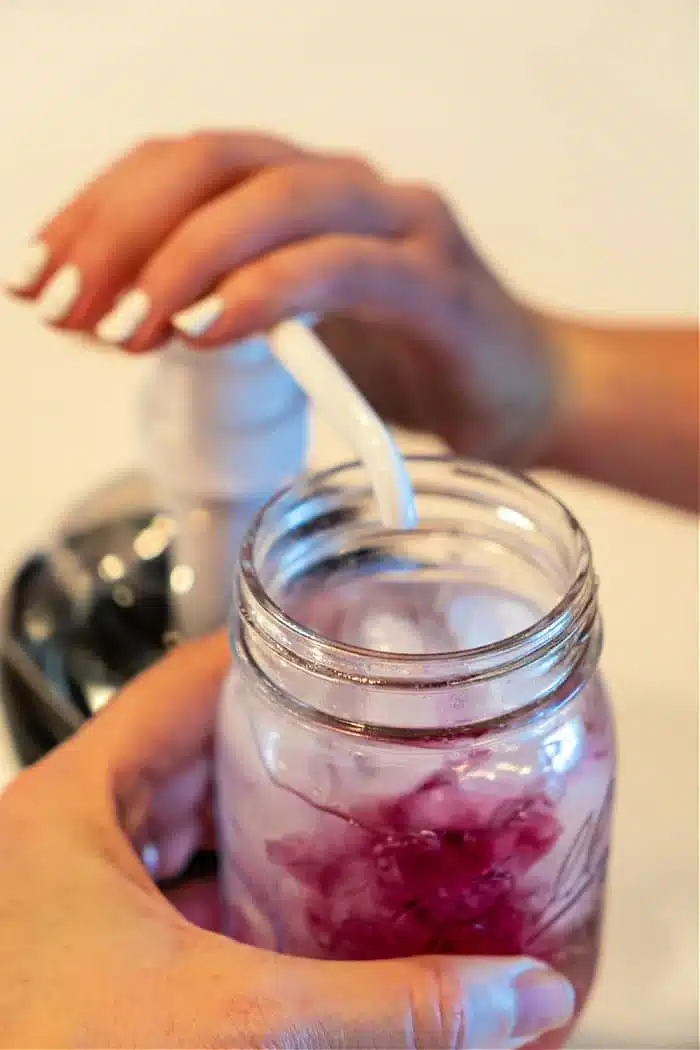 A person pumps liquid from a dispenser into a mason jar filled with a refreshing Lotus iced drink. The person's other hand carefully holds the jar while the pump's nozzle hovers above it, adding the perfect touch. In the softly blurred background, hints of vibrant colors dance subtly out of focus.