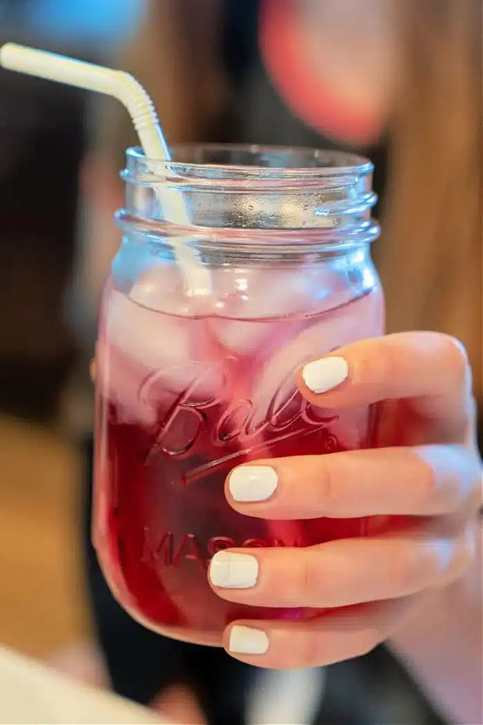 A hand with white-painted nails holds a mason jar filled with a pink iced Lotus drink and a white straw. The jar is half-full, radiating refreshing vibes against the softly blurred background.