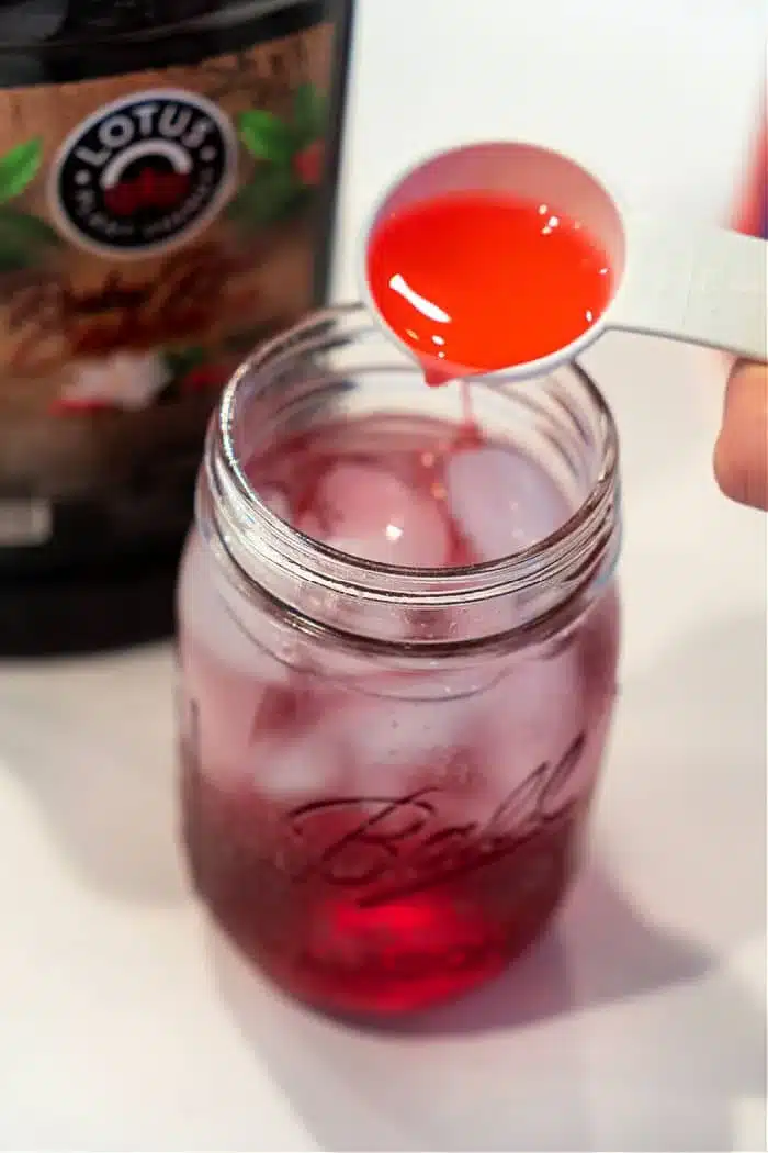 A measuring spoon pours red liquid into a Ball Mason jar packed with ice and another red liquid, creating a tantalizing Lotus Drink. A partially visible bottle labeled Lotus hints at the refreshing flavors waiting to be discovered.