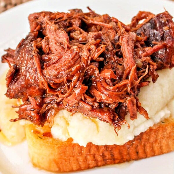 A savory Beef Manhattan graces the table, featuring a slice of toasted bread topped with creamy mashed potatoes and shredded beef, all elegantly presented on a white plate with a woven placemat underneath.