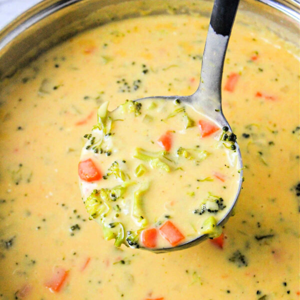 A ladle serving creamy broccoli beer cheese soup filled with broccoli florets and carrot pieces.