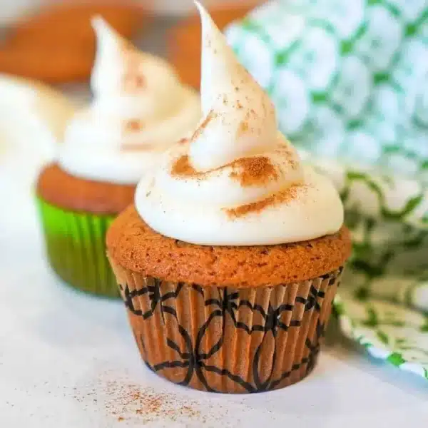 Two gingerbread muffins topped with a swirl of cream and a sprinkle of cinnamon sit on a white surface. The muffins are in decorative wrappers, one green and one with a black pattern. A green and white patterned cloth is partially visible in the background.