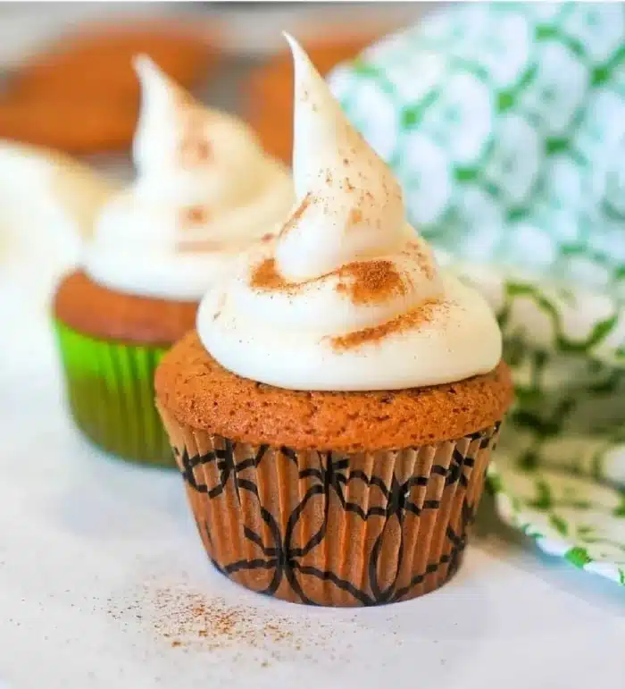 Two gingerbread muffins topped with a swirl of cream and a sprinkle of cinnamon sit on a white surface. The muffins are in decorative wrappers, one green and one with a black pattern. A green and white patterned cloth is partially visible in the background.