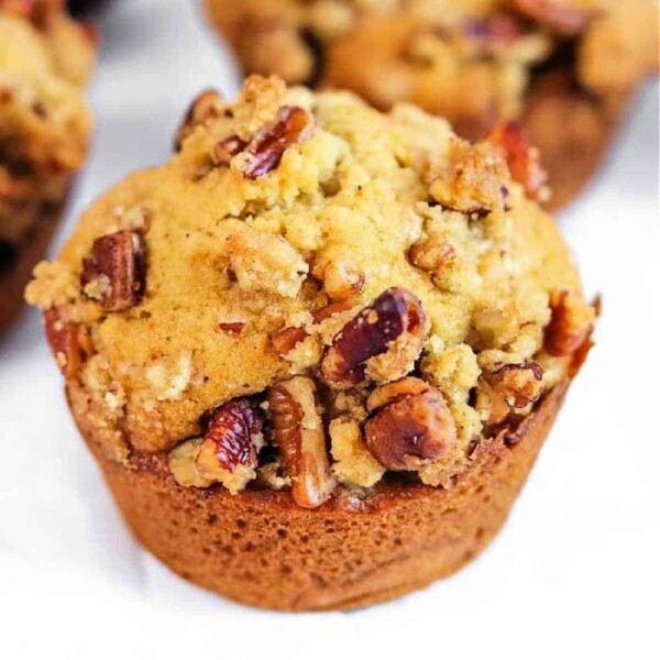 A close-up of a sweet potato muffin with a crumbly top and pecans, set on a light surface.