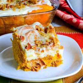 A slice of frosted pumpkin cake topped with chopped nuts is on a white plate, evoking the warmth of pumpkin spice. A tray with more cake is in the background, and a fork rests nearby on a colorful cloth reminiscent of cinnamon rolls.