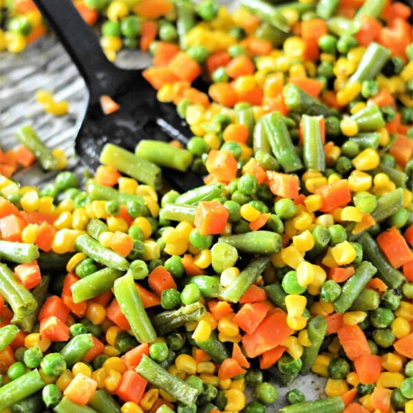 A close-up of a colorful mix of frozen vegetables ready for roasting on a baking sheet, featuring green beans, peas, corn, and diced carrots. A black spoon is partially visible among the vibrant veggies.